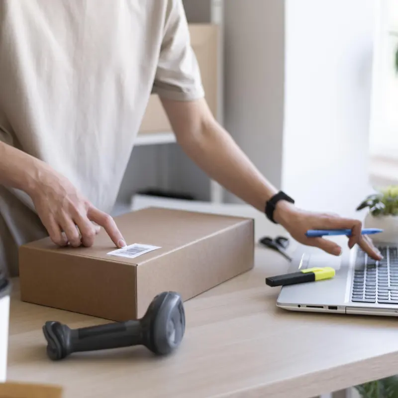 Person an Laptop Eine Person steht an einem Schreibtisch mit einem Laptop, einem Packet und einem Handscanner. Sie tippt etwas in den Laptop ein was sie auf dem Paket abliest